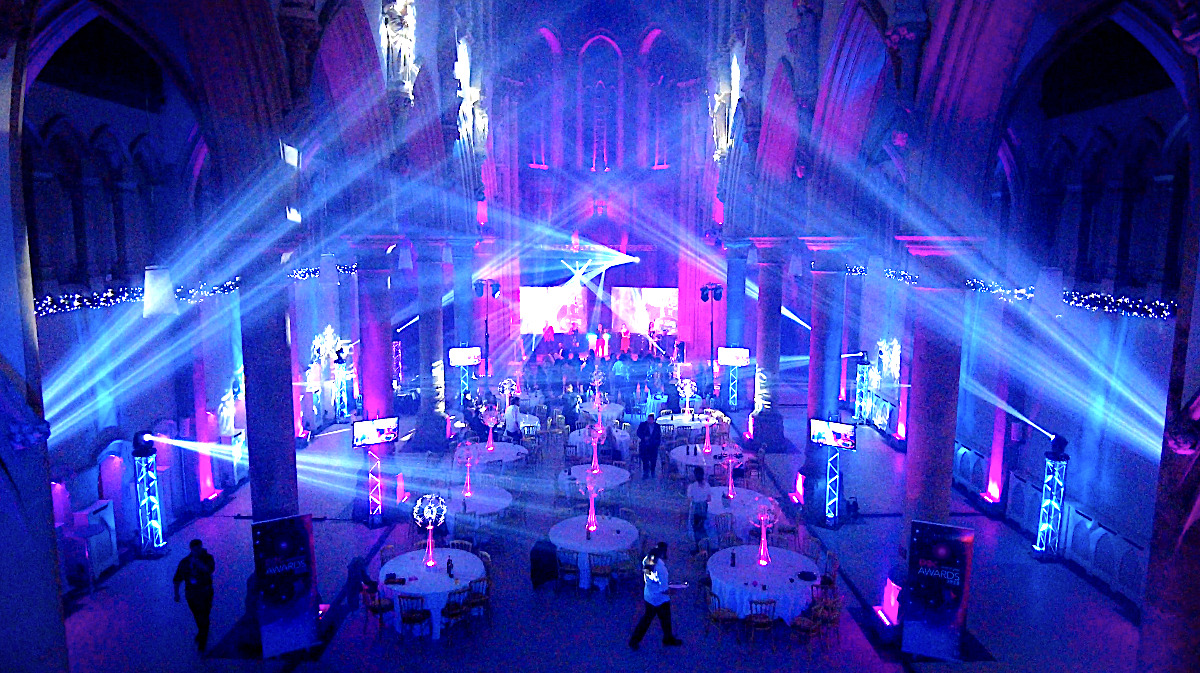 Event lighting image from back of the Gorton monastery manchester with Blue gobo beams from moving lights, there is a stage with two large screens and lighting equipment sat on truss stands with uplights making them shine cyan blue Monastery Manchester Multi Blue beams